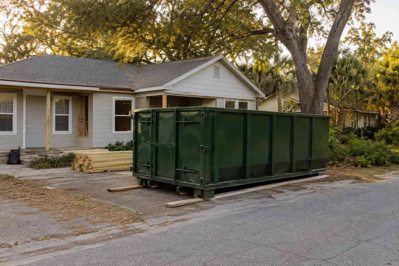 Green roll-off dumpster beside a bungalow under live oaks and palms